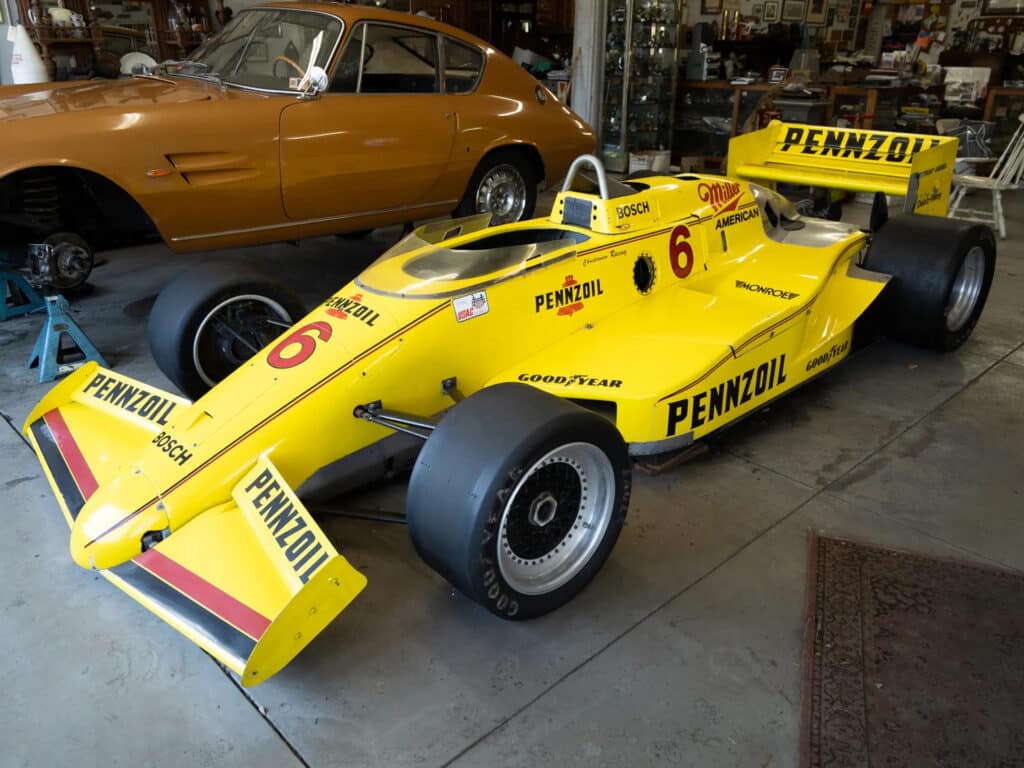 La imagen muestra un coche de carreras vintage, específicamente el March 85C, diseñado por Adrian Newey. El coche está pintado de un color amarillo brillante con la marca “Pennzoil” y el número 6 en su lateral. Tiene grandes neumáticos negros Goodyear y un prominente alerón delantero con rayas rosas y moradas en el borde inferior. El vehículo está estacionado dentro de lo que parece ser un garaje o taller, con varias herramientas y otro coche en el fondo. Este coche representa una pieza de la historia del automovilismo, destacando el trabajo de diseño de Adrian Newey, conocido por sus contribuciones innovadoras en la Fórmula Uno.