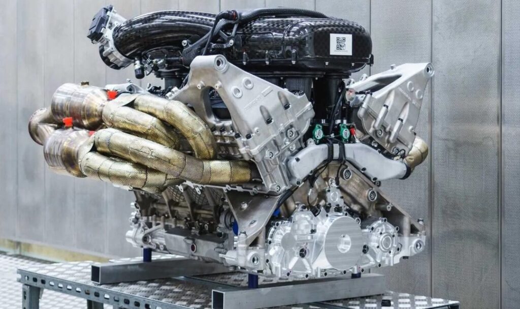 La imagen muestra un motor de alto rendimiento, presumiblemente del Aston Martin Valkyrie, montado en un soporte para exhibición o ensamblaje. El motor es complejo, con numerosos componentes visibles: hay colectores de escape envueltos en material resistente al calor, un colector de admisión de fibra de carbono en la parte superior y varias otras piezas mecánicas de metal. La artesanía y la ingeniería que se han invertido en la construcción de este motor lo hacen interesante, especialmente para aquellos que aprecian la tecnología automotriz y los coches deportivos de alta gama.