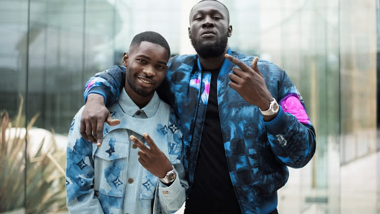 Imagen de dos personas frente a un edificio de cristal. La persona a la izquierda lleva una chaqueta de mezclilla con un patrón de logotipos repetidos, posiblemente de una marca de lujo. La persona a la derecha lleva una chaqueta azul con detalles en rosa brillante, también sugiriendo una etiqueta de diseñador. Ambos visten ropa casual en lugar de trajes formales, lo que ejemplifica el cambio en el tipo de consumidor que compra marcas de lujo, destacando la tendencia actual de preferir ropa deportiva y colorida sobre la vestimenta tradicional.