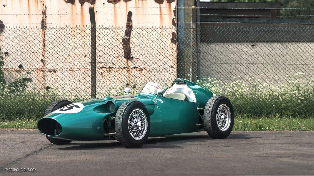 La imagen muestra un Aston Martin DBR5, un coche de carreras vintage, estacionado sobre una superficie de asfalto con un fondo de vegetación y una cerca. El coche está pintado de un color verde brillante y presenta el diseño clásico de ruedas descubiertas típico de los coches de carreras de mediados del siglo XX. Tiene ruedas de radios, una gran parrilla frontal y la carrocería parece estar bien mantenida o restaurada.