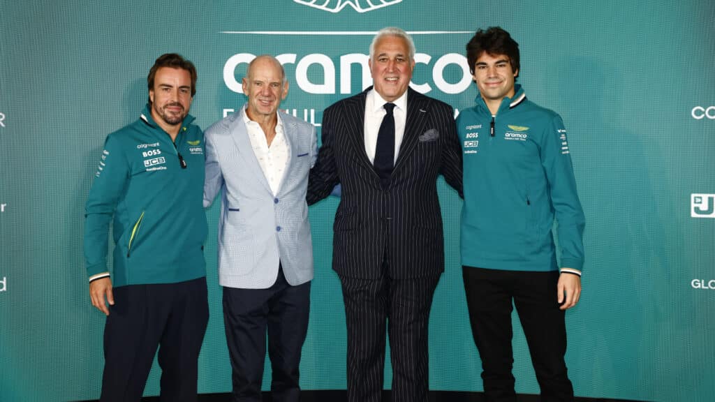 La imagen muestra a cuatro personas de pie frente a un fondo con el texto “Cognizant” visible en la parte superior. De izquierda a derecha, están Fernando Alonso y Adrian Newey, seguidos por Lawrence Stroll y Lance Stroll. Los dos primeros llevan uniformes del equipo con logotipos de patrocinadores, mientras que los dos últimos están vestidos con trajes formales.