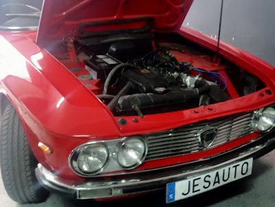 Imagen de la parte frontal de un coche clásico Lancia Fulvia rojo con el capó abierto, mostrando el compartimento del motor. El coche tiene una parrilla distintiva y faros redondos, característicos de los diseños de automóviles clásicos. La matrícula dice “JESAUTO”.