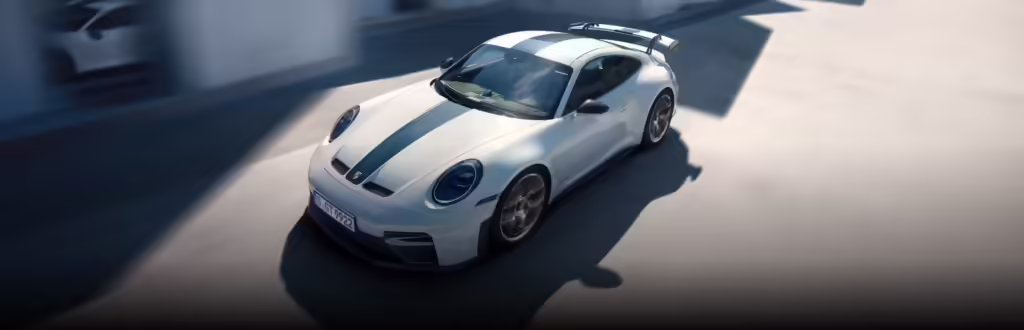 Imagen de un Porsche 911 GT3 blanco con distintivas franjas de carreras, posicionado sobre una superficie lisa con un fondo desenfocado que sugiere movimiento o velocidad. El coche es un modelo de alto rendimiento, asociado con lujo y velocidad, debido a su diseño elegante y el contexto del paquete Weissach, que ofrece mejoras aerodinámicas y reducción de peso.