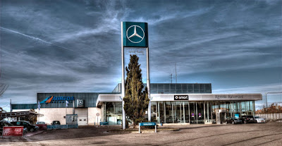 Imagen de un concesionario de Mercedes-Benz durante el día. La fachada del edificio es predominantemente de vidrio con marcos metálicos y el logotipo de Mercedes, una estrella dentro de un círculo, está montado en la parte superior del edificio y en un poste alto frente a él. Hay cielo azul con algunas nubes dispersas arriba y varios coches estacionados alrededor. El nombre ‘Mercedes’ es claramente visible en la parte frontal del edificio.