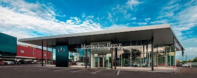 Imagen del exterior de un edificio moderno con grandes ventanas de vidrio y un techo plano, con un cartel prominente que dice “Mercedes-Benz” junto con el logotipo de Mercedes-Benz. El cielo está despejado con pocas nubes y hay un amplio estacionamiento frente al edificio.