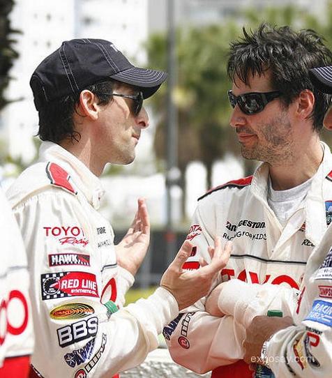 Keanu Reeves (a la derecha) en el día de prensa de la carrera Toyota Pro/Celebrity en 2010. Ambos individuos llevan trajes de carreras con múltiples logotipos de patrocinadores como TOYO TIRES, HAWK, RED LINE y BBS.