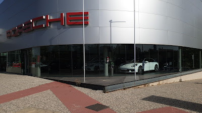 Imagen del exterior de un edificio moderno con ventanas de vidrio reflectante. Sobre la entrada, hay un cartel rojo con letras blancas que dice “Porsche.” Frente a la fachada de vidrio, hay un coche deportivo blanco en exhibición. El entorno sugiere que es un concesionario de coches llamado Artal Automoción.