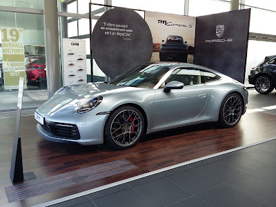 Imagen de un coche Porsche plateado exhibido en un showroom. El coche está posicionado frente a pancartas promocionales, una de las cuales tiene el logotipo de Porsche y menciona “911 Carrera S”. El entorno sugiere que es una exhibición de vehículos de lujo, posiblemente para marketing o propósitos de exhibición, perteneciente a Artal Automoción.