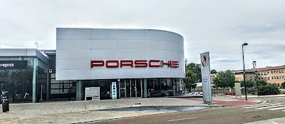 Imagen de un edificio moderno con una fachada curva y grandes ventanas. El nombre “PORSCHE” está claramente visible en el edificio en letras rojas mayúsculas. El entorno sugiere que es un concesionario o showroom de vehículos Porsche, perteneciente a Artal Automoción.