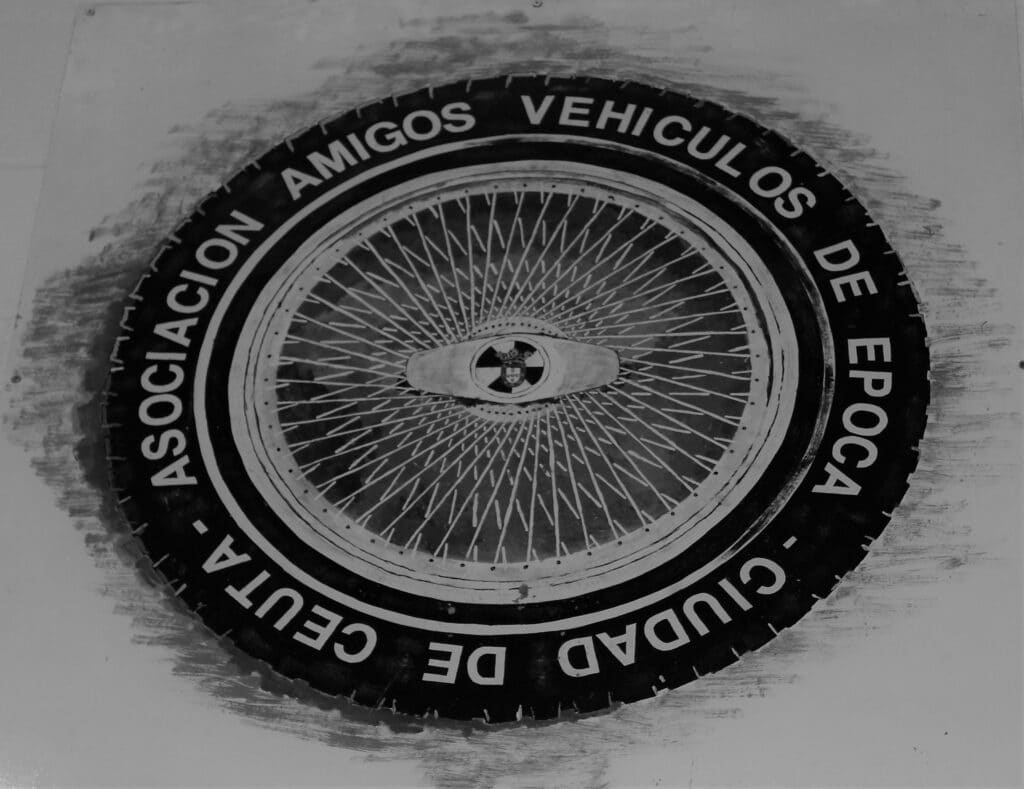 Logotipo de un club de automóviles clásicos. El gráfico central es una rueda con radios y un logotipo en el medio. Alrededor de la rueda, el texto dice ‘ASOCIACIÓN AMIGOS VEHÍCULOS DE ÉPOCA CIUDAD DE CEUTA’.