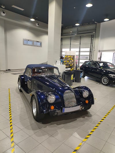 Imagen de un coche clásico con un diseño distintivo, con un largo capó delantero y faros redondeados, estacionado dentro de lo que parece ser un showroom o garaje. El vehículo es predominantemente de color oscuro con detalles cromados. Hay otros coches y algunos equipos en el fondo, así como cinta de precaución amarilla creando un límite alrededor del área donde se exhibe el coche.