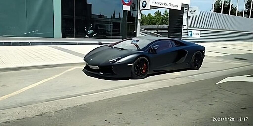 Imagen de un coche deportivo negro estacionado al lado de una calle. El coche tiene un diseño aerodinámico y está situado frente a lo que parece ser un concesionario con paredes de vidrio y el logotipo “Augusta Aragón” visible. El entorno sugiere un ambiente urbano y hay una señal de tráfico que indica prohibido el paso a la izquierda de la carretera.