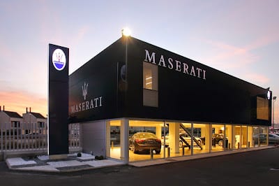 Imagen del exterior de un concesionario de Maserati al anochecer. El edificio es moderno, con grandes ventanas de vidrio que permiten ver el showroom donde se exhibe un coche Maserati. El concesionario tiene dos pisos y está bien iluminado, con el logo de Maserati destacado en la señalización sobre el edificio.
