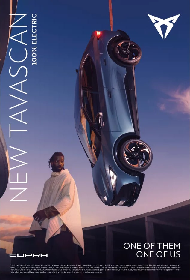 Anuncio del ‘NEW TAVASCAN - 100% ELECTRIC’ de CUPRA. La imagen muestra un SUV eléctrico azul suspendido en el aire contra un cielo crepuscular. Debajo del coche, hay una persona de espaldas al espectador, vistiendo lo que parece ser una capa o abrigo. El texto en la parte inferior dice ‘ONE OF THEM ONE OF US’.