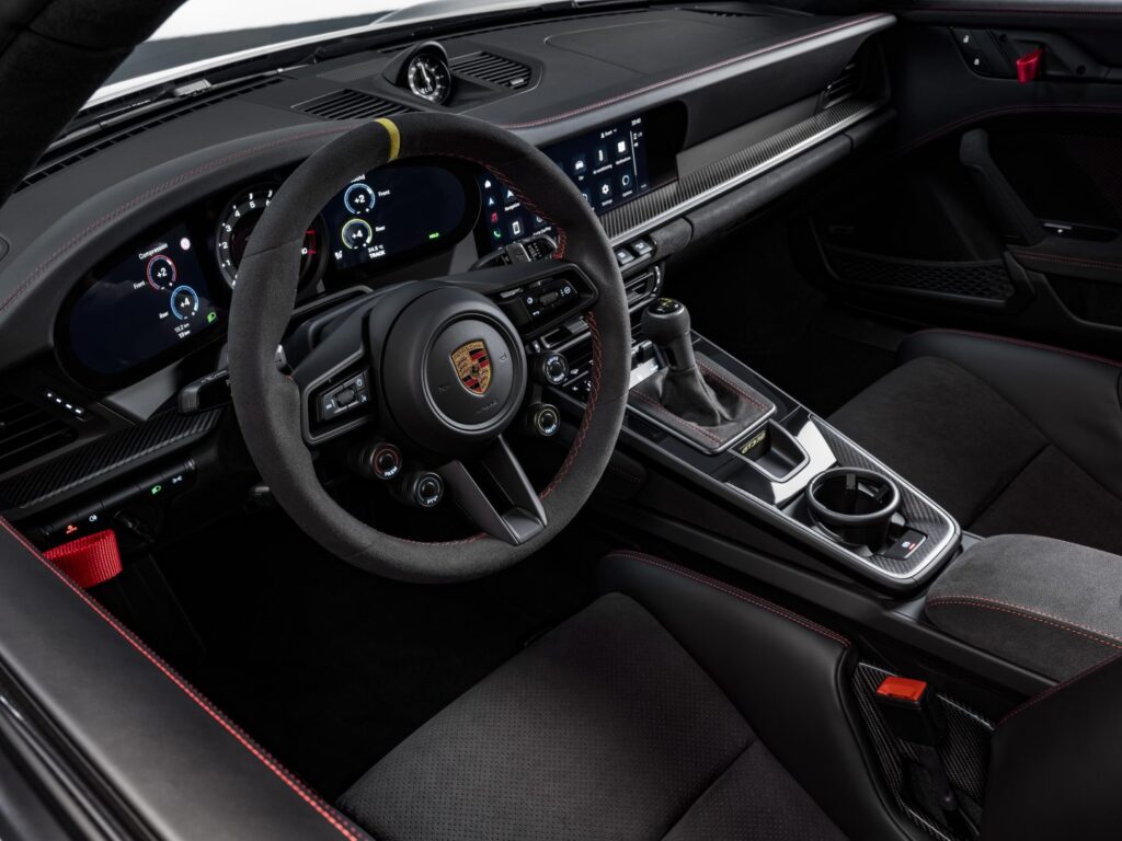Imagen del interior de un Porsche GT3, enfocada en el área del conductor. Se observa un volante con el emblema de Porsche en el centro, múltiples botones de control y levas de cambio detrás. El tablero incluye pantallas digitales con varios indicadores y medidores. La consola central tiene palancas de cambio, interruptores de modos de conducción y otros controles del vehículo. Los asientos tienen detalles y costuras rojas que contrastan con el interior negro.