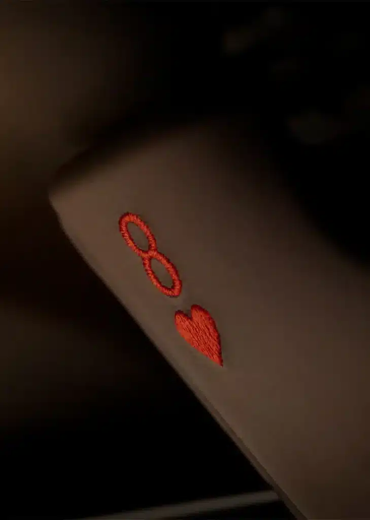 Vista de cerca de una superficie de cuero oscuro con dos símbolos bordados: un signo de infinito y un corazón, ambos en hilo rojo. El bordado es detallado, mostrando la textura de las puntadas. Esta imagen representa un detalle del interior del Aston Martin DB12 Goldfinger Edition, indicando personalización o marca.