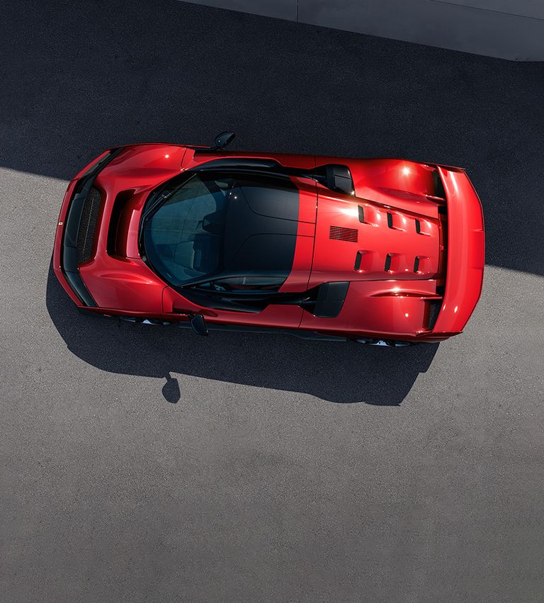 Vista superior de un Ferrari F80 rojo estacionado sobre una superficie gris. El coche presenta un diseño aerodinámico con líneas elegantes, y el techo parece estar hecho de vidrio u otro material transparente, ofreciendo una vista del interior. La sombra del coche es visible en el suelo, indicando que la iluminación proviene de arriba.