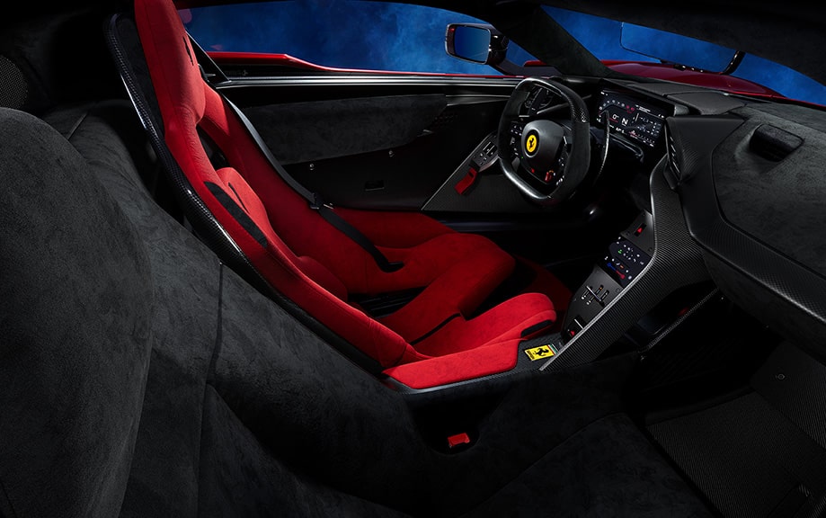 Interior de un Ferrari F80, centrado en el lado del conductor. El interior es lujoso, con asientos deportivos rojos que tienen un prominente logo del caballo encabritado, emblema de Ferrari. El tablero y el volante son negros con detalles en rojo, y hay varias pantallas digitales visibles. El diseño es elegante y moderno, enfatizando el rendimiento y el lujo típicos de la marca.
