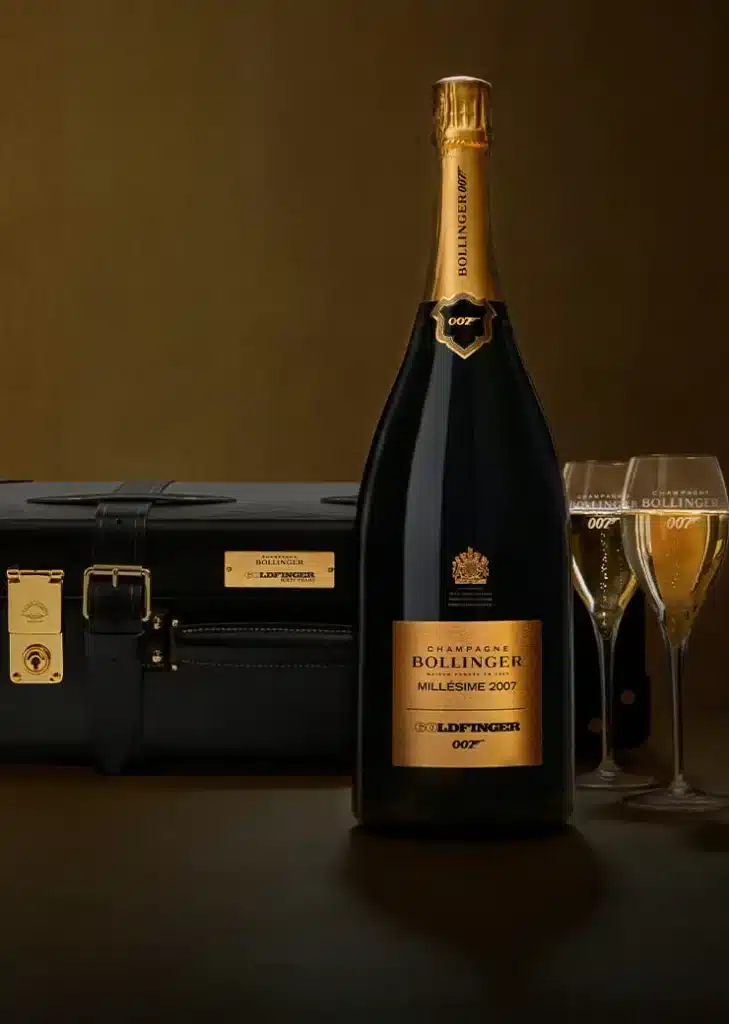 Botella de Champagne Bollinger Millésime 2007 junto a dos copas de champagne y un maletín negro con cerraduras doradas. El maletín tiene el número ‘007’, indicando una conexión con James Bond. Esta imagen sugiere un tema de sofisticación y lujo, asociado con el Aston Martin DB12 Goldfinger Edition.