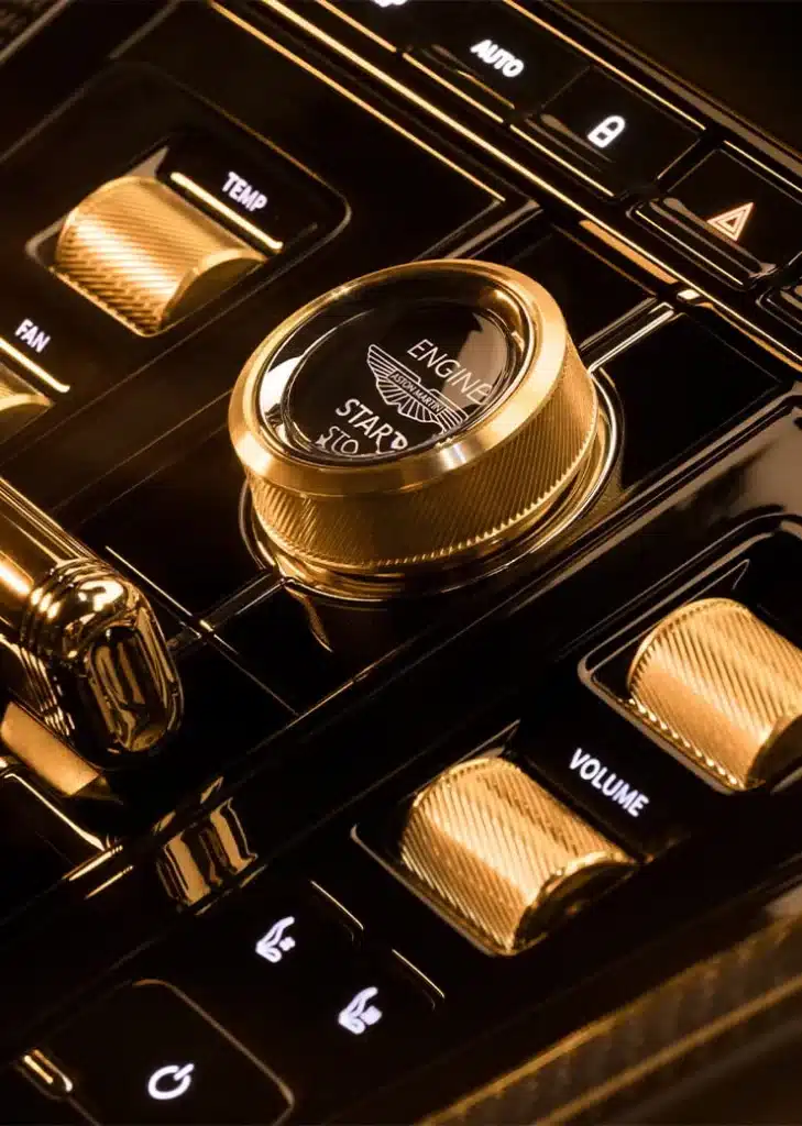 Primer plano del interior de un Aston Martin DB12 Goldfinger Edition, enfocando la consola central con varios controles. El elemento más destacado es un botón dorado de encendido/apagado del motor, etiquetado ‘ENGINE START STOP’. Alrededor de este botón hay otros botones negros con símbolos blancos, incluyendo uno para ajustar el volumen, sugiriendo que esta área se utiliza para gestionar tanto el encendido del vehículo como el sistema multimedia.