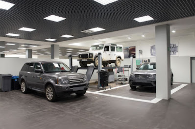 Imagen del interior del taller de Land Rover de Automóviles Sánchez. Se ven tres vehículos: un Land Rover Discovery negro en el suelo, un Land Rover Defender más antiguo elevado en un elevador de coches en el centro, y otro Land Rover Discovery negro en el suelo a la derecha. El taller está limpio y bien organizado, con un patrón de suelo ajedrezado, iluminación brillante y paneles acústicos en el techo.