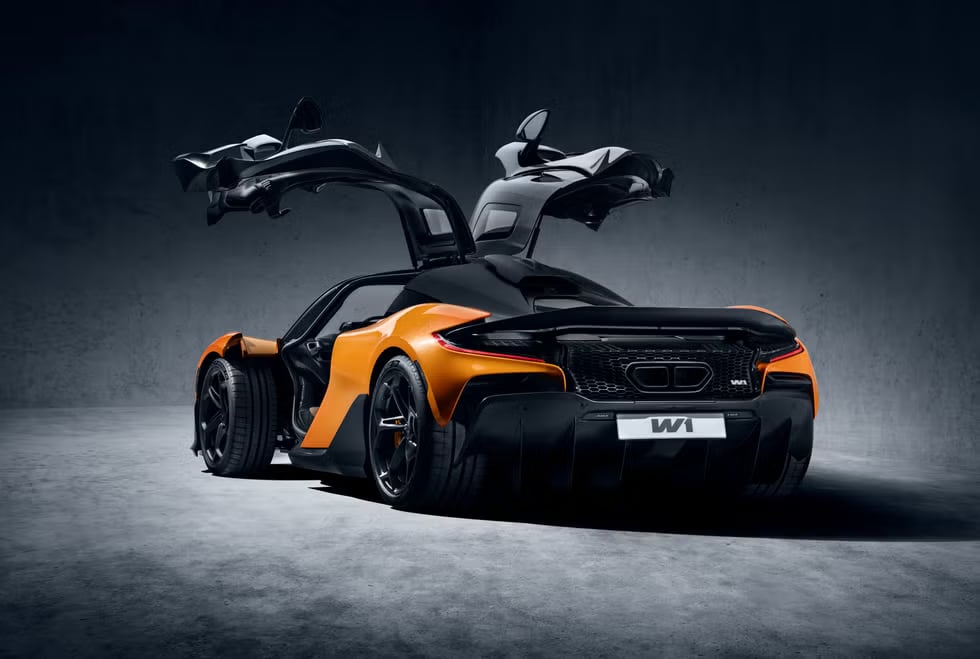 Vista lateral y trasera de un McLaren W1 de color naranja y negro con las puertas abiertas hacia arriba. El coche está posicionado de manera que se destacan su perfil lateral y trasero, enfatizando su diseño y características aerodinámicas.