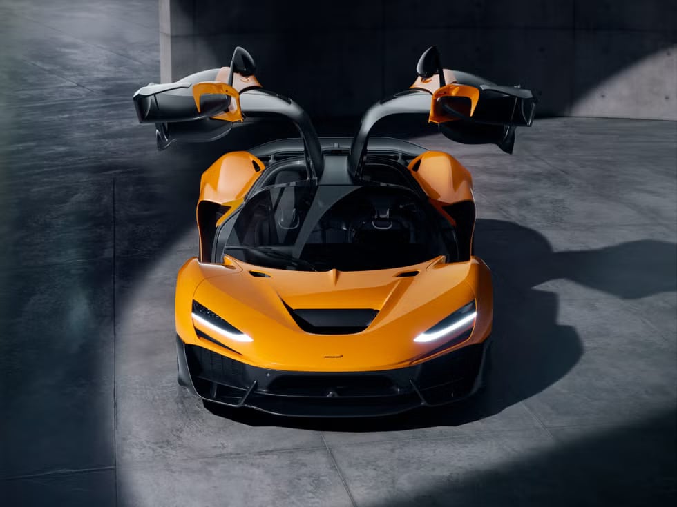 Automóvil deportivo McLaren W1 de color naranja con las puertas abiertas hacia arriba en estilo mariposa. El vehículo está estacionado dentro de una estructura de concreto con poca iluminación, posiblemente un garaje o sala de exposición. El diseño del coche es elegante y aerodinámico, con faros distintivos que le dan un aspecto agresivo.
