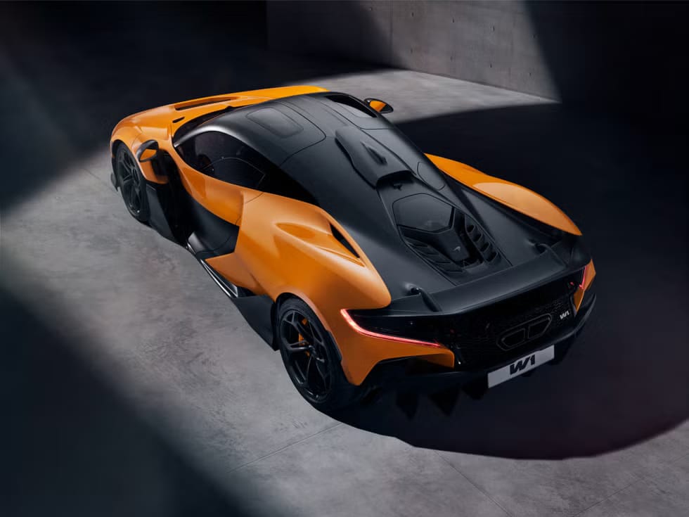 Vista superior de un automóvil deportivo McLaren W1 de color naranja con detalles en negro, estacionado en un garaje o sala de exposición con poca iluminación. El diseño aerodinámico del coche y sus líneas elegantes se destacan bajo la luz.