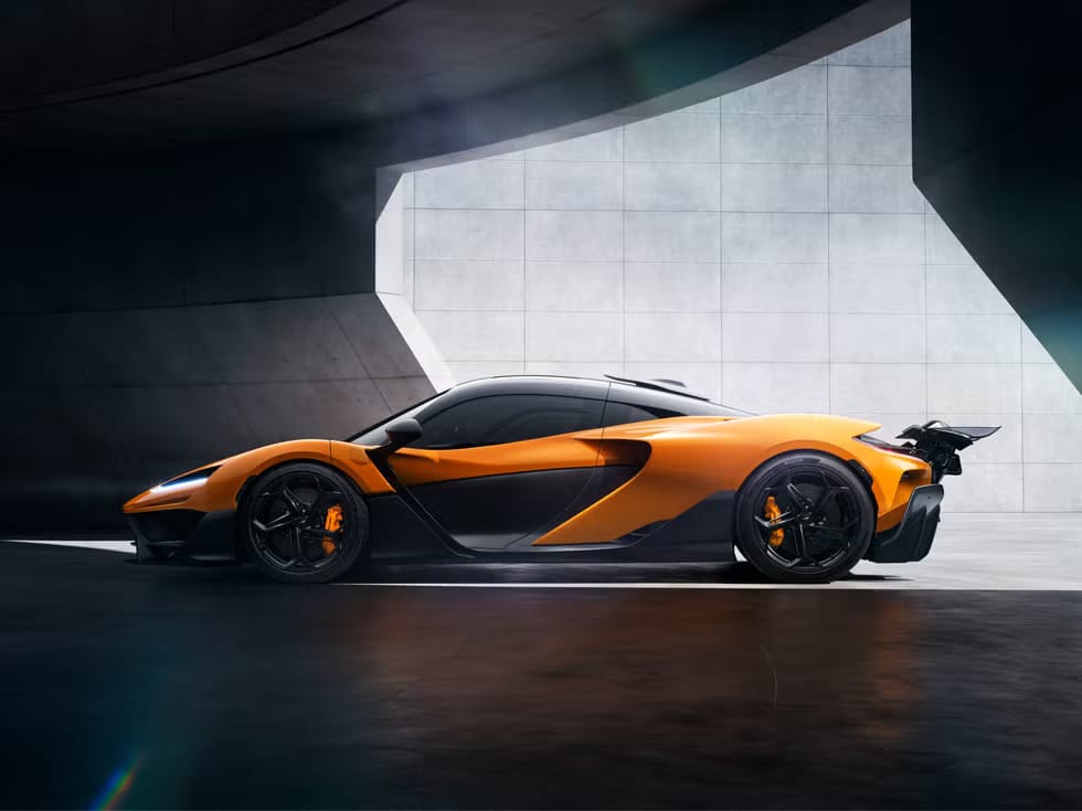 Vista de un McLaren W1 de alto rendimiento con un esquema de colores naranja y negro. El coche está posicionado en un ángulo que resalta su diseño aerodinámico, incluyendo un alerón trasero prominente y líneas de carrocería elegantes. El vehículo está situado en un entorno minimalista con superficies de concreto liso, lo que contrasta con el estilo agresivo del coche.