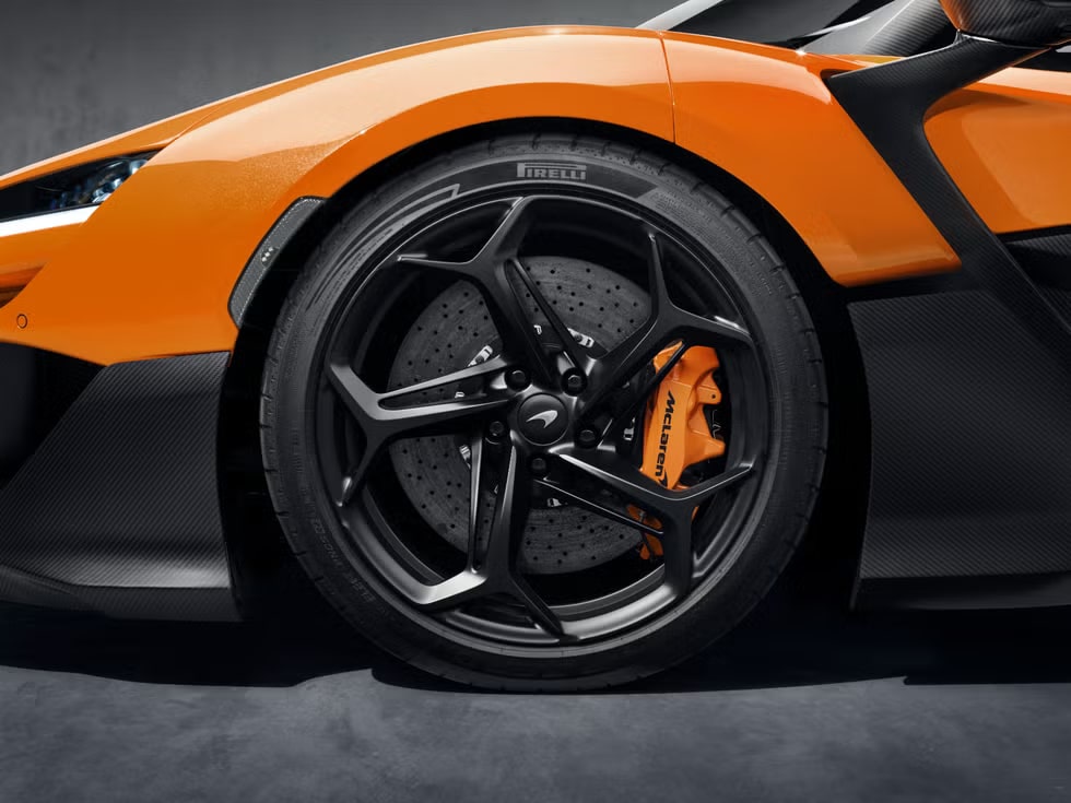 Vista en primer plano de la rueda de un McLaren W1 de color naranja, destacando el diseño intrincado del rin negro y el sistema de frenos de alto rendimiento con una pinza de freno naranja. El logo de Pirelli en el neumático indica una marca conocida por sus neumáticos de alto rendimiento.