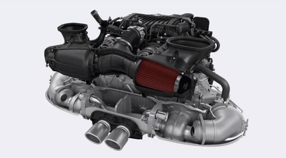 Imagen del motor del nuevo Porsche 911 GT3. Se observan los detalles intrincados de los componentes del motor, incluyendo el sistema de admisión de aire con un filtro rojo, cubiertas de plástico negro en la parte superior y tubos de escape metálicos en la parte inferior. El motor parece ser una configuración de seis cilindros opuestos, característica de los vehículos de alto rendimiento de Porsche. El fondo es blanco, lo que resalta el diseño y la complejidad del motor.