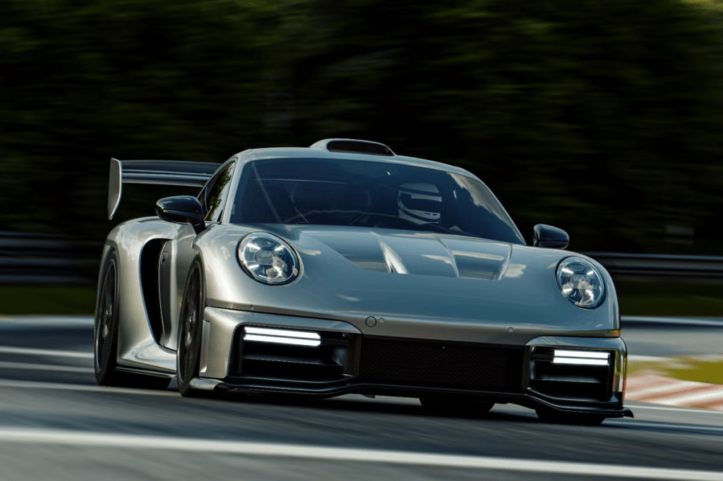 Un Porsche 911 RML P39 plateado en movimiento en una pista de carreras, con elementos aerodinámicos distintivos como un gran alerón trasero, un divisor frontal y guardabarros ventilados. Los faros del Porsche están encendidos y parece estar en una situación de conducción dinámica con árboles borrosos en el fondo, indicando movimiento rápido.