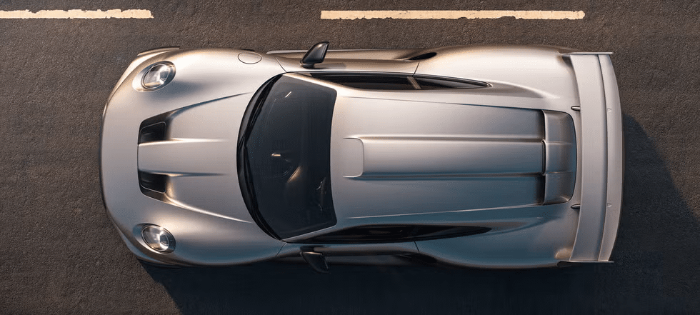 Vista superior de un Porsche 911 RML P39 plateado estacionado sobre una superficie de asfalto con marcas viales. El coche tiene un diseño aerodinámico con curvas elegantes y dos tomas de aire visibles en el capó, indicando que es un vehículo de alto rendimiento.