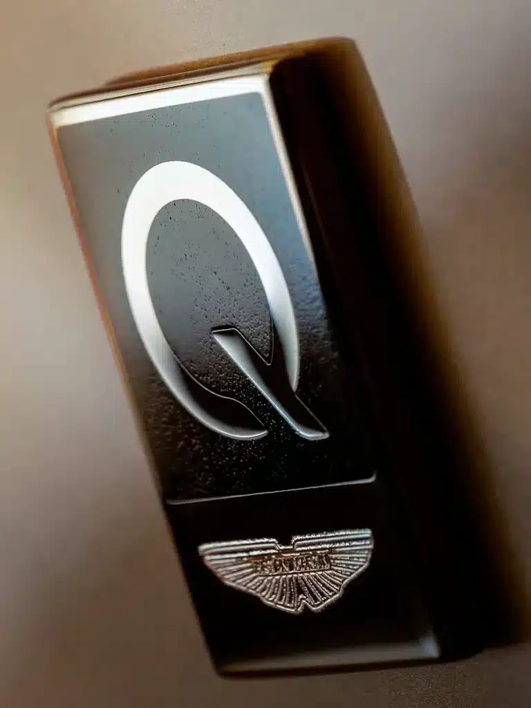 Primer plano de un objeto metálico con una superficie reflectante, que presenta un símbolo en relieve o grabado que parece ser una letra ‘Q’ estilizada con un diseño de hoja en su interior. El fondo está desenfocado con tonos degradados de claro a oscuro, sugiriendo que el objeto está iluminado desde un lado, posiblemente en un entorno interior.