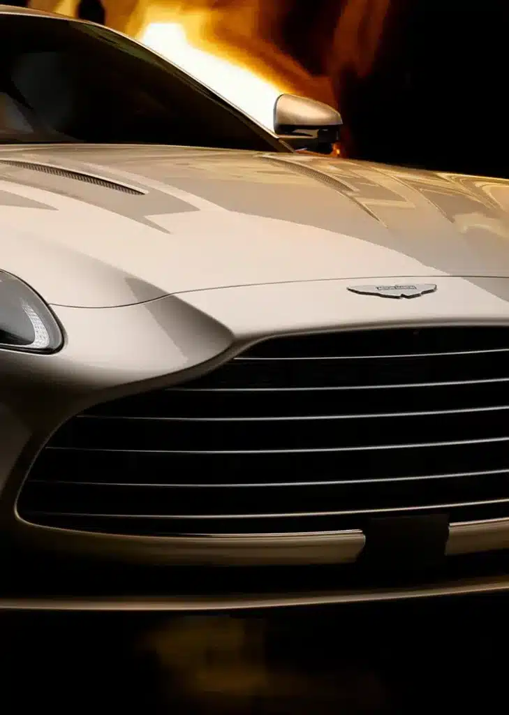 Vista frontal de un Aston Martin DB12 Goldfinger Edition. El vehículo presenta un diseño elegante con la icónica parrilla y el emblema de Aston Martin en la parte delantera. El coche es de un color claro, posiblemente plateado o dorado claro, que complementa su nombre ‘Goldfinger’. El fondo tiene un resplandor cálido que acentúa la elegancia y el estatus de alta gama del coche.