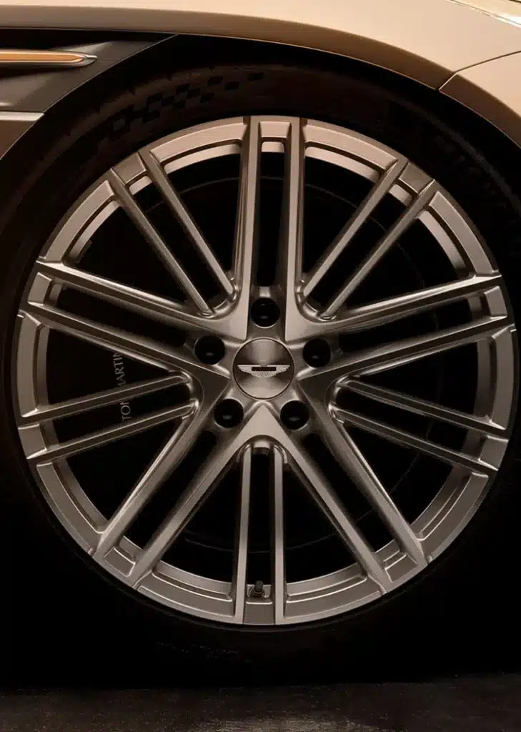 Vista de cerca de la rueda de un Aston Martin DB12 Goldfinger Edition. La rueda tiene un diseño de múltiples radios y el centro lleva el emblema de Aston Martin con las icónicas alas. El neumático muestra patrones de banda de rodadura visibles, indicando que está diseñado para uso en carretera.