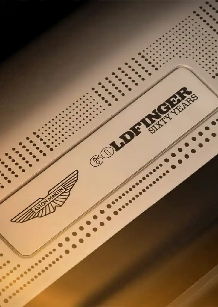 Primer plano de un objeto con el texto ‘GOLDFINGER’ y ‘SIXTY YEARS’ impreso. Debajo del texto ‘SIXTY YEARS’, hay un emblema con un diseño de alas estilizadas. El fondo tiene un patrón de puntos y el tono general es dorado y negro, sugiriendo que podría ser un artículo conmemorativo relacionado con el 60 aniversario de ‘Goldfinger’.