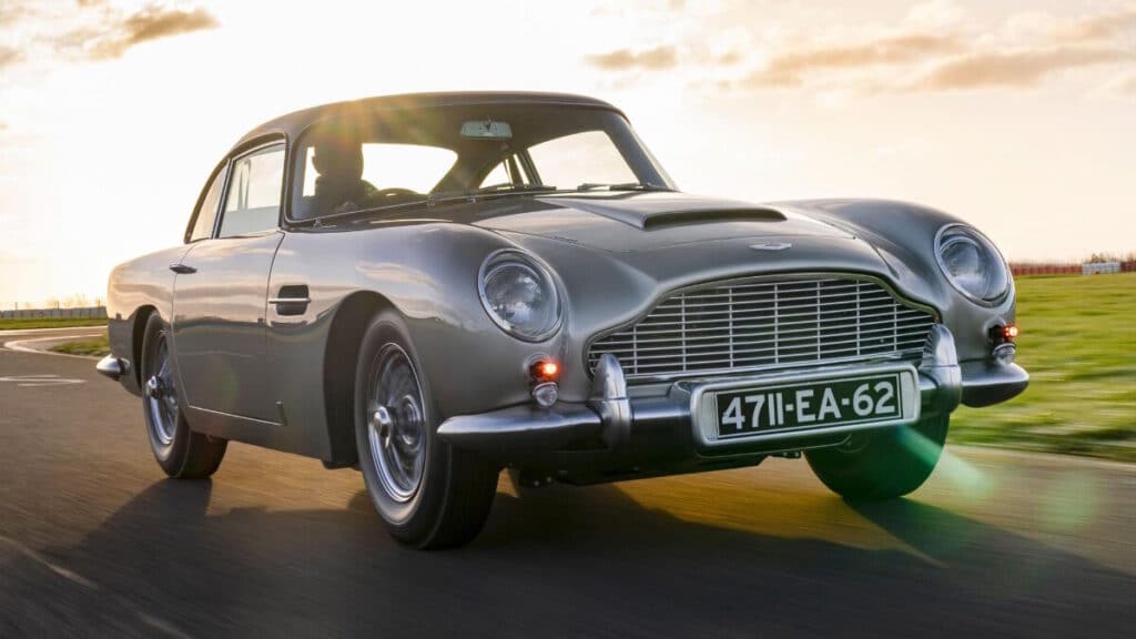 Imagen de un automóvil Aston Martin clásico de color gris, en movimiento sobre una carretera con un fondo desenfocado que muestra el cielo al atardecer y campos verdes. El vehículo tiene la placa ‘471-EA-62’ y presenta un diseño elegante y retro con faros redondos y una parrilla frontal distintiva.
