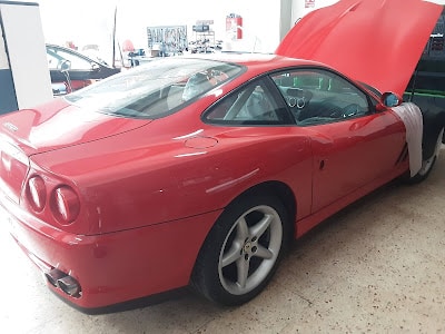 Imagen de un automóvil deportivo Ferrari de los años 90, de color rojo con la capota abierta, mostrando el motor. El vehículo tiene dos puertas y está estacionado en un interior, posiblemente un showroom o taller, con otros vehículos y objetos al fondo. Destaca por su diseño aerodinámico y su color llamativo.