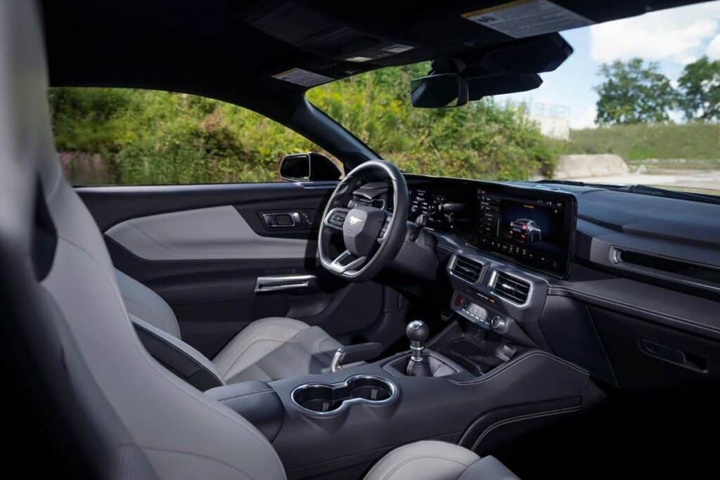 Vista interior del Ford Mustang Raptor 2026, mostrando asientos de cuero negro con un diseño moderno. El tablero presenta un panel táctil horizontal y elegante que se extiende hacia el lado del pasajero. Debajo, hay rejillas de ventilación y controles de clima. El volante tiene un diseño de fondo plano con controles integrados, y la consola central incluye una palanca de cambios y portavasos. La estética general es lujosa y deportiva.