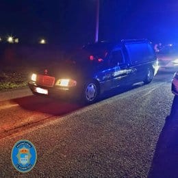 Un coche fúnebre Mercedes-Benz detenido en O Porriño por la Guardia Civil al lado de una carretera de noche, con las luces delanteras y de emergencia encendidas. Hay otros vehículos con las luces encendidas en el fondo, sugiriendo tráfico activo. El coche fúnebre fue detenido por participar en una carrera ilegal.