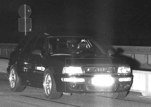 Imagen en blanco y negro de un Audi RS2 en movimiento de noche. En el asiento del conductor hay una figura que se asemeja al Monstruo de las Galletas de Sesame Street, con ojos saltones y pelaje azul. El vehículo está pasando frente a un letrero circular con borde rojo y fondo blanco, típico de señales de tráfico prohibido.
