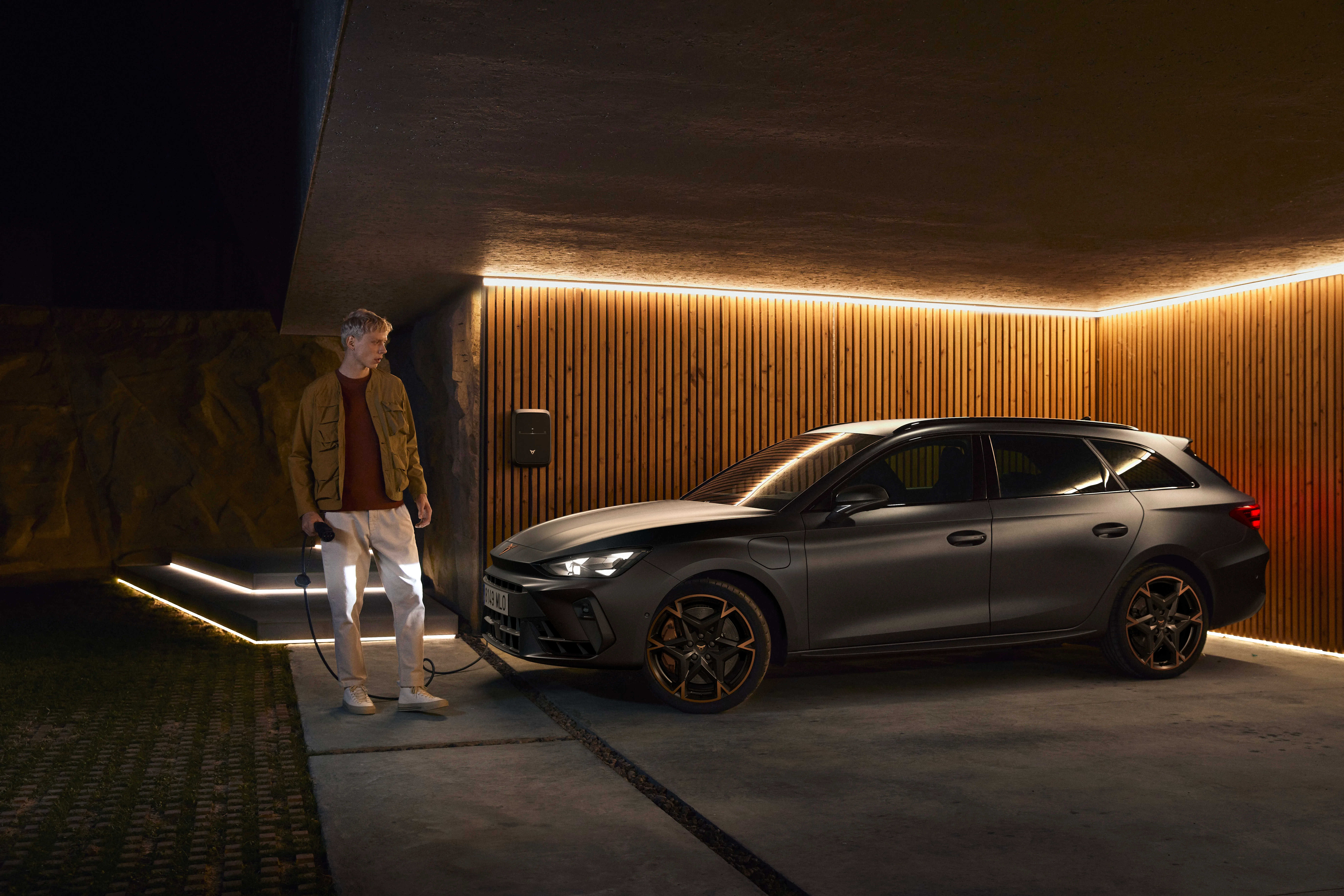 Imagen de un automóvil Cupra estacionado bajo una estructura con iluminación ambiental cálida. El vehículo es de color oscuro y presenta un diseño deportivo con rines detallados y faros encendidos. Junto al coche, hay una figura humana con ropa casual, parada y mirando hacia el vehículo. La escena tiene lugar en un entorno nocturno que resalta la iluminación del coche y la arquitectura circundante.