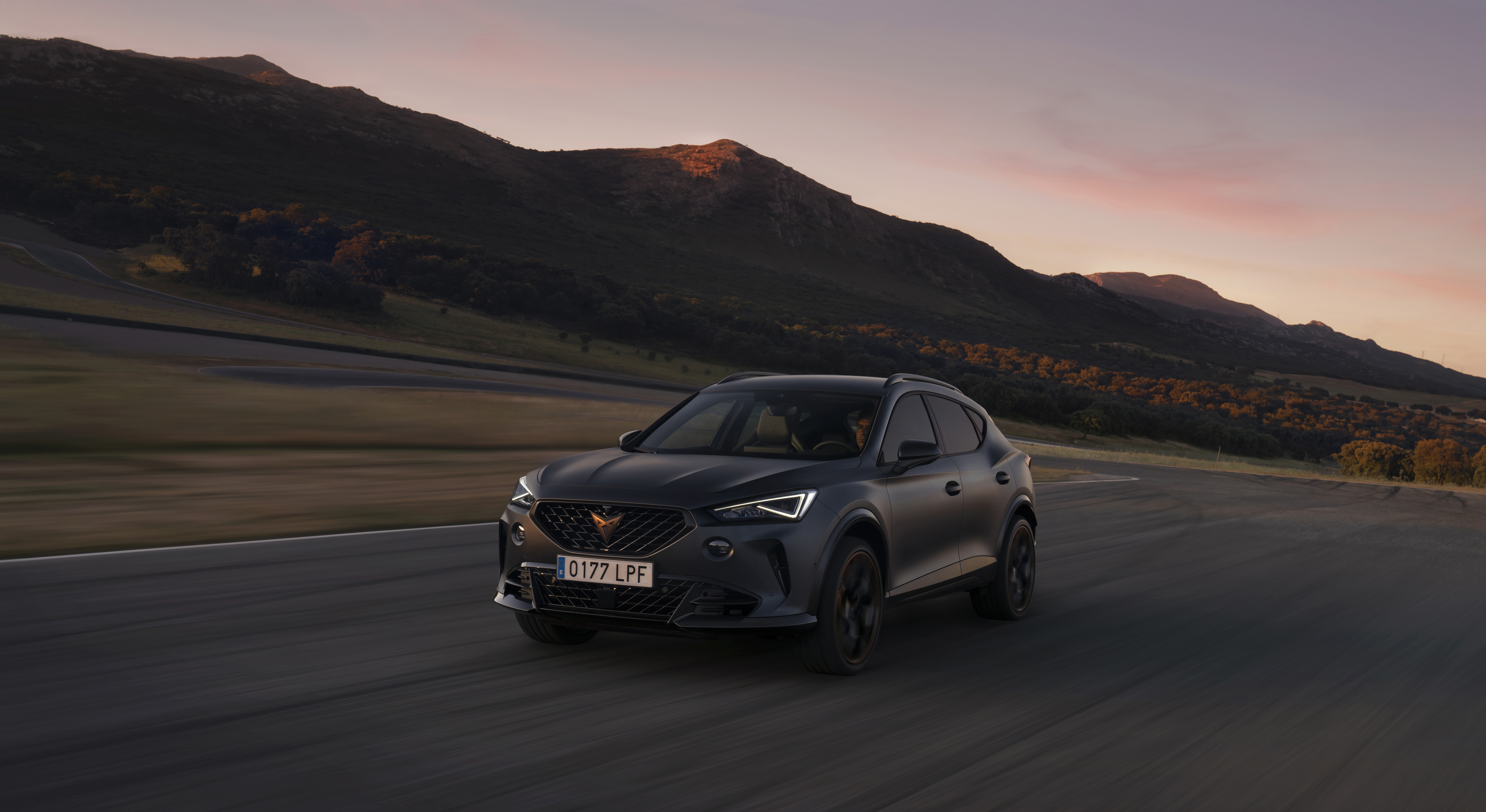 Imagen de un CUPRA Tavascan conduciendo por una carretera con montañas al fondo durante el amanecer o atardecer. Las condiciones de iluminación crean un efecto dramático, destacando las características de diseño del vehículo, como su forma aerodinámica, la parrilla frontal distintiva y los faros. La matrícula del coche es visible con la inscripción ‘0127JFP’.