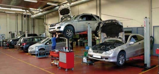 Imagen del interior del taller Mercedes-Benz de Ágreda Automóvil. Se ven varios coches, presumiblemente modelos de Mercedes-Benz, en diferentes estados de reparación o mantenimiento. Algunos vehículos están elevados en elevadores con los capós abiertos, indicando que se está trabajando en los motores. Al menos un técnico es visible trabajando en un coche. El taller está equipado con herramientas y maquinaria típicas para el servicio de vehículos.