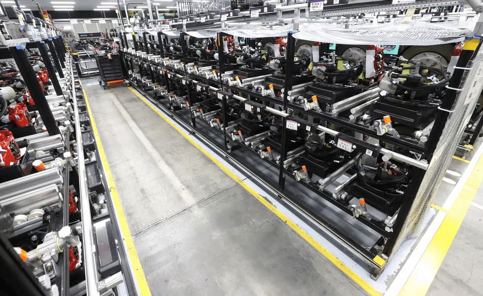 Imagen de una línea de ensamblaje en una fábrica, con múltiples filas de maquinaria y piezas de automóviles, probablemente para ensamblar vehículos. La maquinaria es predominantemente negra y metálica, con elementos rojos que podrían ser herramientas o accesorios. El suelo tiene marcas de seguridad amarillas que delimitan pasillos y áreas de trabajo.