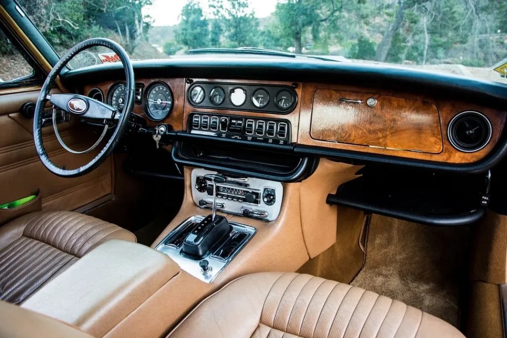 La imagen muestra el interior de un Jaguar XJ6 de los años 70, destacando los acabados de lujo y la alta calidad típicos de este coche clásico. El tablero combina madera y cuero, con una variedad de indicadores y controles. El volante es grande y tiene un diseño clásico, y los asientos están tapizados en cuero beige con un patrón acanalado. La consola central incluye una palanca de cambios y varios controles, enfatizando la atención al detalle y la artesanía del coche. Esta imagen es interesante y relevante porque muestra la elegancia y sofisticación de los vehículos Jaguar vintage, conocidos por sus interiores lujosos y materiales de alta calidad.