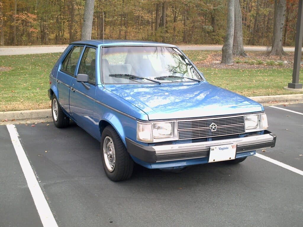 La imagen muestra un Dodge Omni azul estacionado en un aparcamiento. El coche tiene una matrícula de Virginia y parece ser un modelo antiguo, posiblemente de la década de 1980. El coche está estacionado en un espacio designado con líneas blancas que marcan los límites. El fondo incluye una zona de césped con árboles y una carretera. El diseño del coche presenta una forma cuadrada con faros rectangulares y una parrilla cromada. La imagen es interesante porque captura un vehículo vintage en buen estado, lo que podría atraer a los entusiastas de los coches o a aquellos interesados en la historia automotriz.