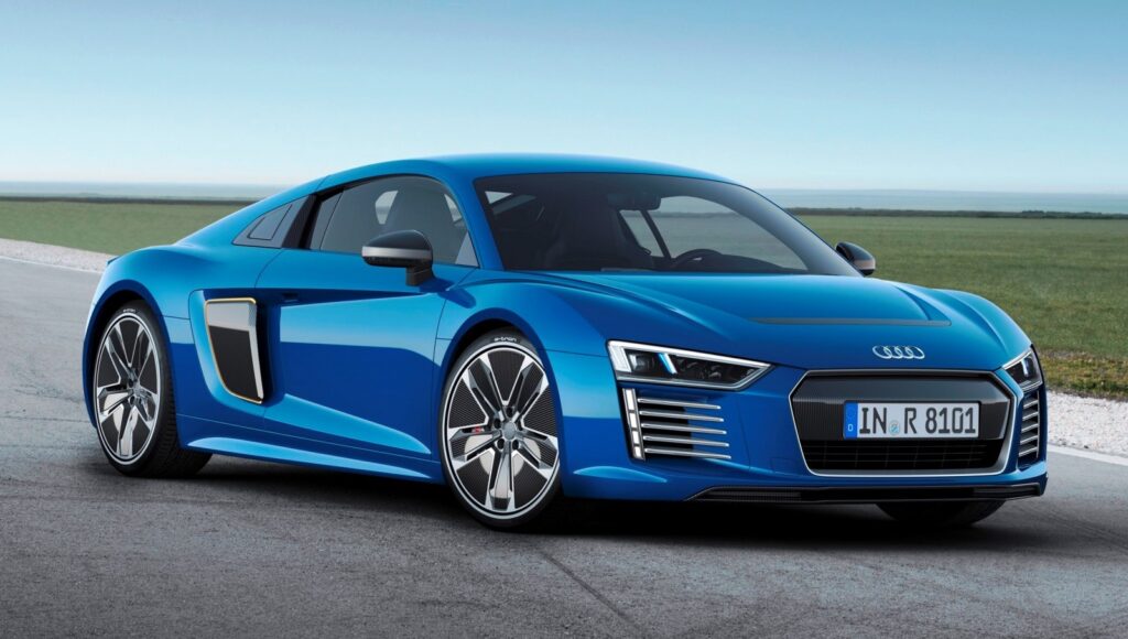 La imagen muestra un Audi R8 e-tron azul estacionado en una carretera con un campo de hierba y un cielo despejado en el fondo. El coche presenta líneas aerodinámicas elegantes, faros LED distintivos y una parrilla frontal prominente con el logotipo de Audi. La matrícula dice "IN R 8101". Esta imagen es interesante y relevante porque destaca el diseño y el atractivo estético de un vehículo de lujo de alto rendimiento, resaltando sus características modernas y deportivas.