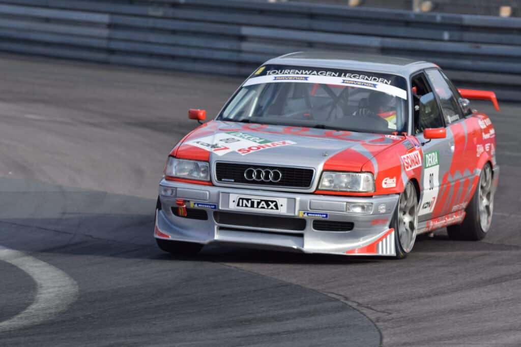 Claro, aquí tienes: La imagen muestra un Audi 80 Competition Quattro en una carrera del DTM, pilotado por Frank Biela. El coche está decorado con los colores y patrocinadores típicos de la competición, incluyendo una combinación de rojo, blanco y negro. El coche está en movimiento, capturado en una curva del circuito, con las ruedas delanteras giradas y el fondo ligeramente desenfocado, lo que transmite la velocidad y dinamismo de la escena. El número del coche es visible en la puerta, y se pueden ver otros coches de competición en el fondo. Esta imagen es interesante porque muestra un momento de acción en una carrera histórica del DTM, destacando tanto el coche como el piloto.